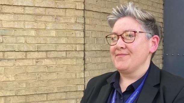 Helen Tebbit with short fair hair wearing brown-rimmed glasses and a dark suit and dark, open-necked shirt. She has a blue lanyard around her neck that has the words "Cambridgeshire Constabulary" written on it. She is standing on the left side of the photograph and looking to her right. Behind her is a light-brown brick wall.

