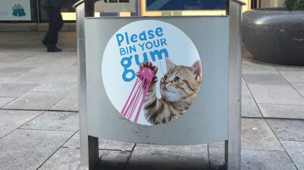 A bin on an urban street. A round sign on the side of the bin has a picture of a cat with some pink gum stuck to its paw and reads: "Please bin your gum".