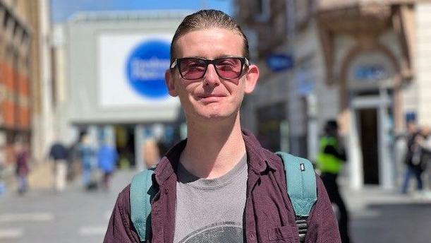 A man wearing dark sunglasses stares into the camera. He is wearing a t-shirt underneath a purple shirt jacket, which is unbuttoned and open, and has an aqua-blue coloured backpack. He is standing in a street, behind him is an entrance to a shopping centre and other shops in the background, which are all out of focus.
