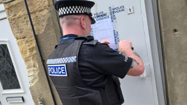The back of a police officer sticking a closure order to the white door of the house.