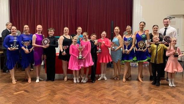 Various ballroom dancers in bright outfits holding trophies.