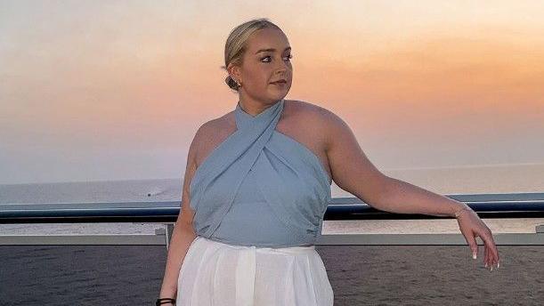 Jessica Thorpe looking at the distance while leaning back on a parapet at sunset. The beach and the sea are behind her. She is wearing a blue top and white trousers and her hair is pulled back. 