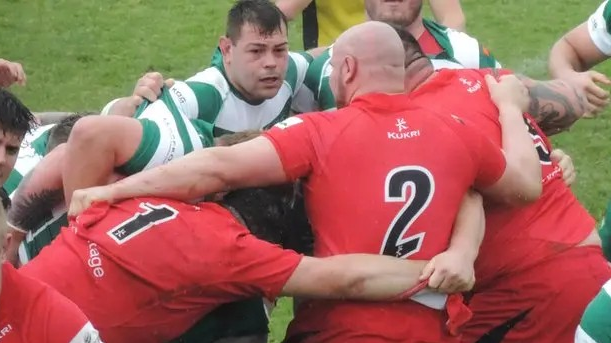 A dark-haired man in a green and white hooped rugby top prepares to go into a scrum with his teammates, against a team wearing red.