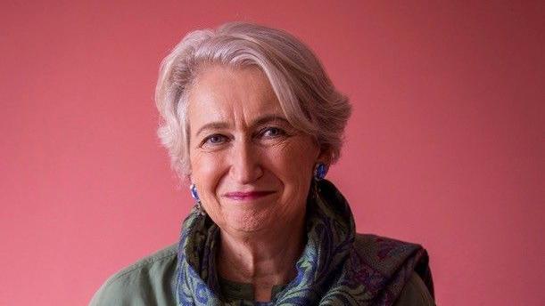 A woman is pictured in front of a pink background with short white hair. She is wearing a green and blue scarf and a green jacket.