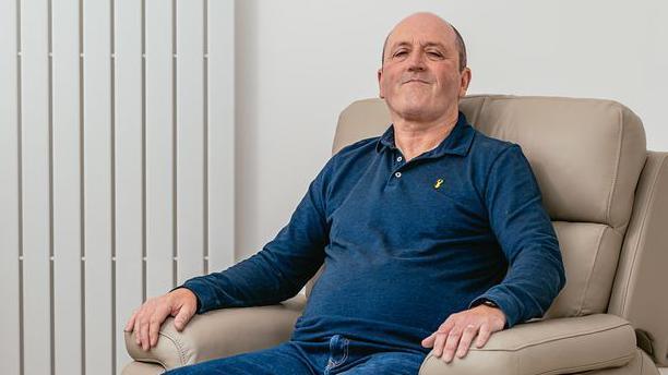 Mike Thomas, who is wearing a blue polo top, is sitting in a cream-coloured armchair and smiling while looking at the camera