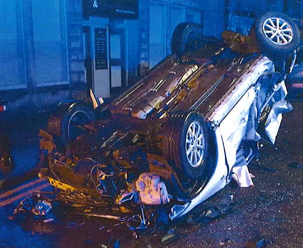 A silver car lying on its roof on the road. It is night time and the detail is unclear. The car is badly damaged. 