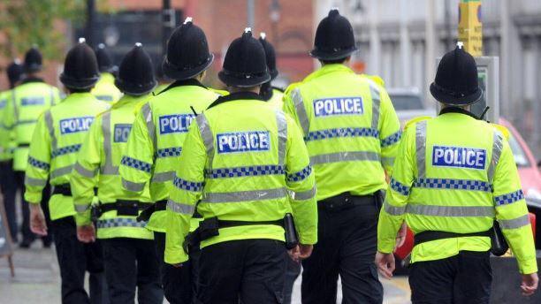 A row of police officers walking down a street. They are wearing traditional police helmets and hi-visibility jackets with blue and silver lettering spelling the word "police".