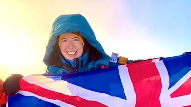 Adriana Brownlee stands and smiles at the peak of a mountain in a blue snowsuit and yellow hat holding a Union Jack flag, with a sunrise behind her