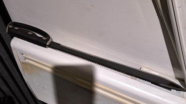 Long straight knife with a pointed end and serrated back upper edge and a black handle with finger guard. It is resting on the top of a white radiator or heater.