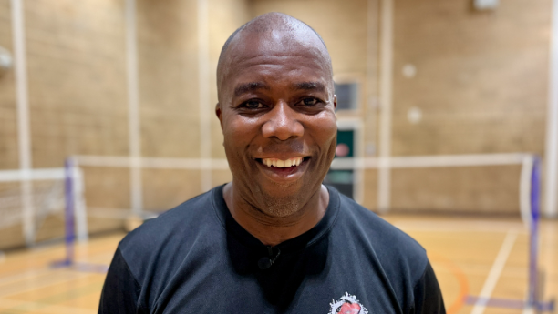 Patrick Lawrence is in the middle of the image standing on a badminton court smiling. He is wearing a navy blue T-shirt.