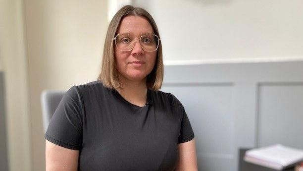 Woman with shoulder length hair and glasses looking seriously in the camera while sat a desk