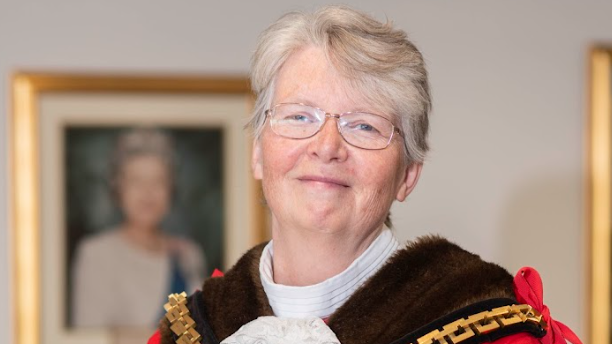 Claire Darke in a portrait. She is wearing glasses and smiling. She has her mayoral dress on and there is a blurred picture of the queen behind her. 