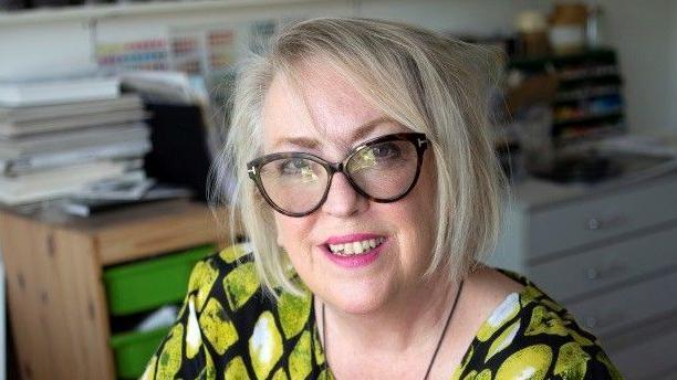 A woman with short blonde hair and large black-framed glasses. She is sitting down wearing a dress with yellow patterns. In the background there are cabinets with folders on top.