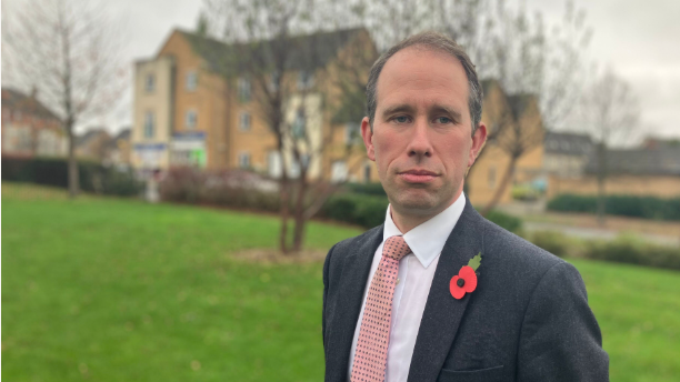 Matthew Barber is pictured on the Great Western Park estate in Didcot. He is standing in front of grass and trees and a small parade of shops can be seen in the background. He is wearing a grey suit, a salmon-coloured tie and a red poppy. 