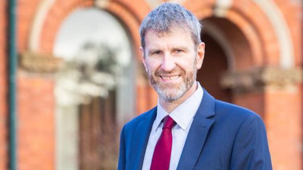 A man with a beard smiling at the camera. He is in front of a red brick building and is wearing a suit and tie