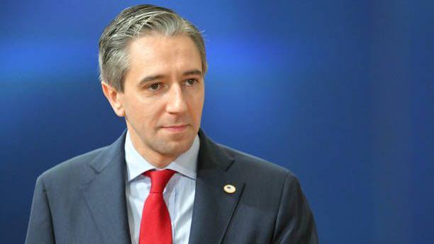 Taoiseach Simon Harris looking sideways at camera in a blue jacket, white shirt and red tie with a blue background behind him