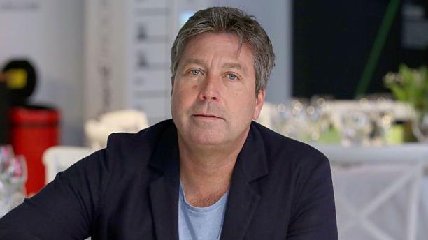 John Torode sat at a dining table, wearing a suit. 