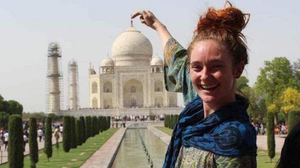 A young woman is posing in front of the Taj Mahal. She's standing down the lawn that leads up to the entrance with her finger pointed at the top of the building. She has wavy auburn hair tied up in bun and is wearing a green top with a blue and black scarf over her shoulders. Small thin trees are lined up along a fountain leading up to the building. The ivory coloured building has rotundas on the roof.