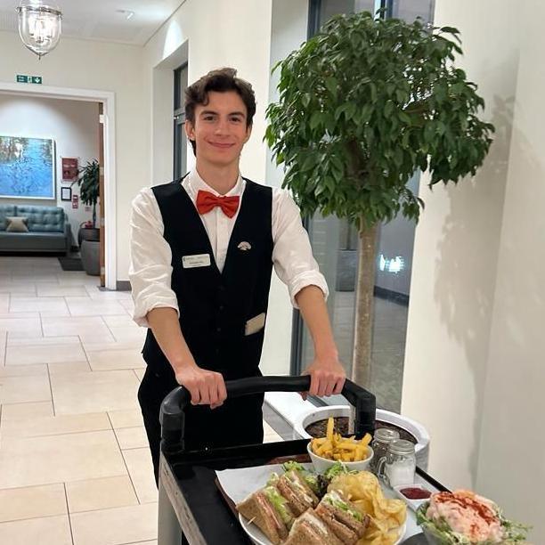 Fernando is dressed in a white shirt and black waistcoat. He has a red bow tie. He is pushing a black trolley with food on it.