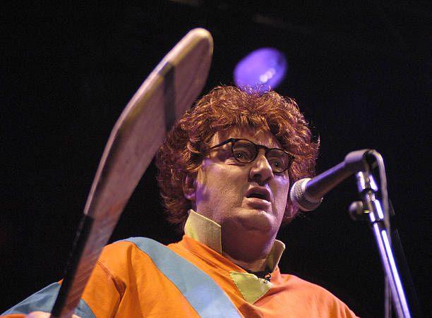 Jon Kenny wearing glasses on stage wearing an orange stripe jacket,  talking into the microphone and holding a hurling bat 