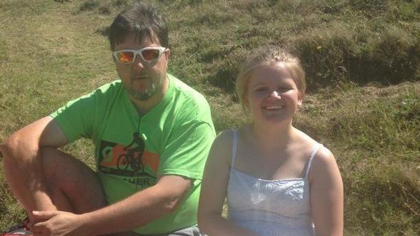 Eve, a young teenage girl wearing a white dress and a ponytail, sits on a grassy hill in the sunshine with her father Nigel, who is wearing a green t shirt and sunglasses