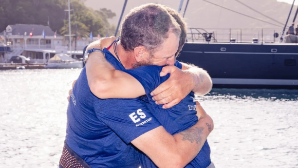 Karl Austen and Ed Shaw wearing navy t-shirts hugging each other on the jetty at English Harbour in Antigua.