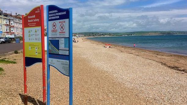 Beach safety signs at the top of the beach at Greenhill, with a street to the left and blue sea and coastal hills in the background