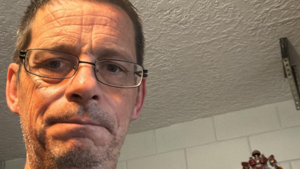 Mr Hartill is wearing glasses in the image which is a close-up selfie. He has brown hair and is in a room with a white ceiling and white tiles below.