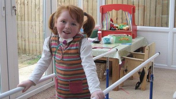 Sophie, with red hair in bunches and wearing a striped dress with a white long-sleeved T-shirt, stands holding white physiotherapy training bars. She has a tube coming from her throat and is smiling at the camera. In the background is a play table with a puppet theatre, next to a child's chair. She is in a conservatory.