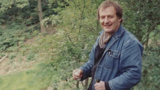 A photo of a man in a denim jacket wearing a scarf and holding a bottle of champagne and a glass. 