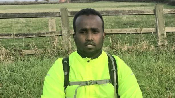 Kasim Ali wear a yellow jacket and sits in front of a fence and grass