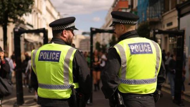 Image of two uniformed police officers stood with their backs to the cameras as people enter gates to Notting Hill Carnival in 2024