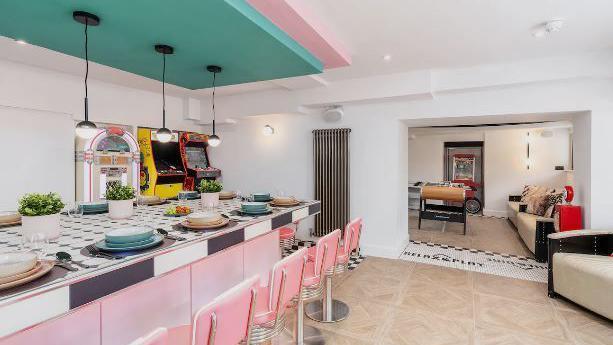 The American-style diner room and games room. There is a large island-style table, with a black and white chequered top and baby pink sides and chairs. The room also has leather sofas, arcade style video games, a juke box and table football.