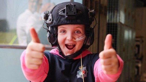 Oliver is pictured smiling wearing a pink and black shirt. He is holding both of his thumbs up.