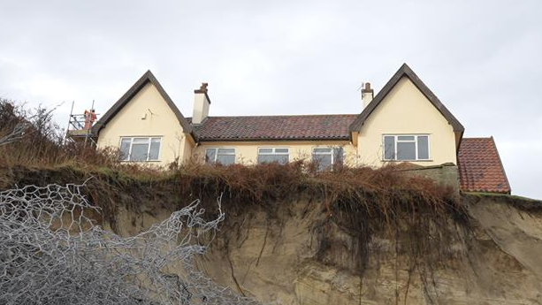 A view from the bottom of a cliff looking up toward a house that can be seen towering over. It is a yellow house with several windows and two chimneys. Only the top storey of the house can be seen. Metal wires can be seen toward the bottom left of the image as part of sea defences. 