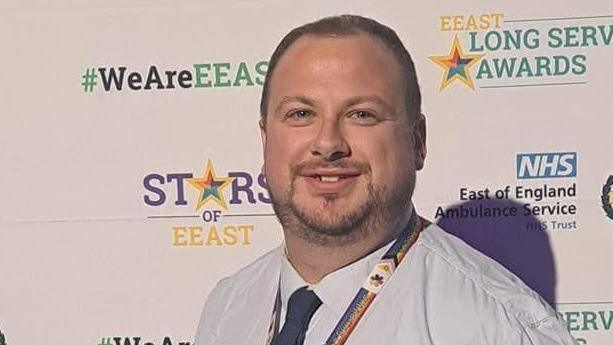 Ben Cronin smiles at the camera during an awards event. He has very short dark hair and has a short beard. He wears a shirt with a blue tie and lanyard around his neck. He stands against a white backdrop that has various businesses logos on it.