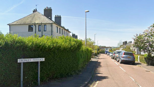A residential street with cars parked and a hedge outside the houses. A street sign saying Dronley Avenue is in the foreground