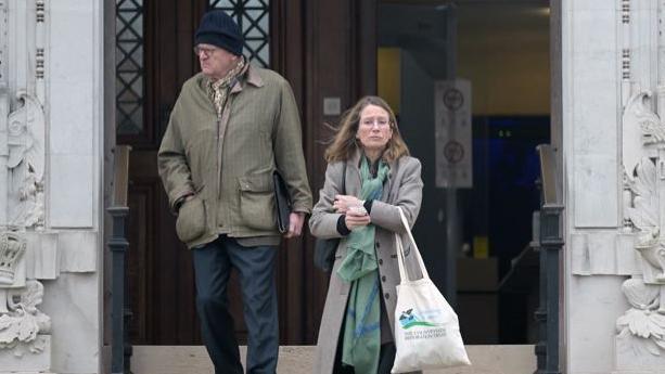 A woman and a man coming down steps outside Newport Crown Court. The man, on the left, wears dark blue trousers and a khaki coat, and a dark blue woolly hat. The woman, on the righ, wears a grey coat and green patterned scarf; she carries a tote bag.