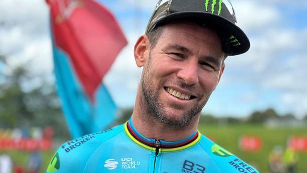 Mark Cavendish smiles at the camera. He is wearing a blue cycling top and a black cap.