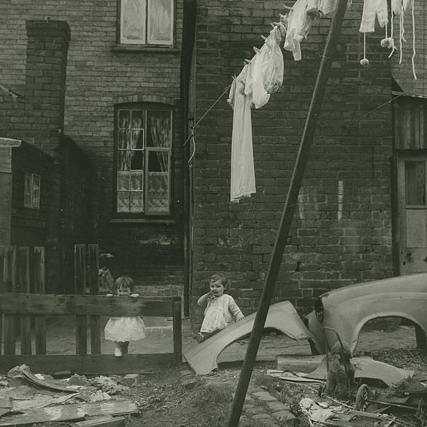 An image of two young girls stood outside an old terraced house with a washing line hanging a number of white items of clothing