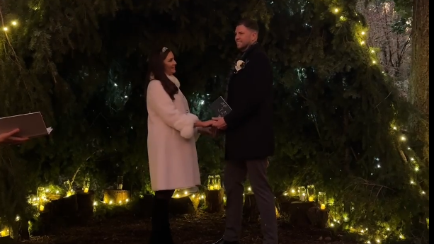 Sinead and Michael hold hands during their wedding ceremony which took place in a forest in front of an array of conifer branches and fairy lights