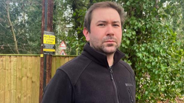 Callum Ringer looks at the camera. He has a serious expression. He has a short beard and short brown hair. He is wearing a black zip-up fleece and is standing outside with a fence, hedge and electricity pole behind him.