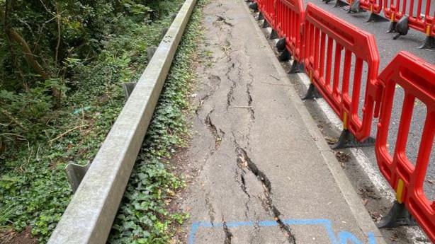 Cracking in the footpath can be seen alongside the A36. The path is uneven. Red traffic bollard can be seen between the bath and the carriageway.