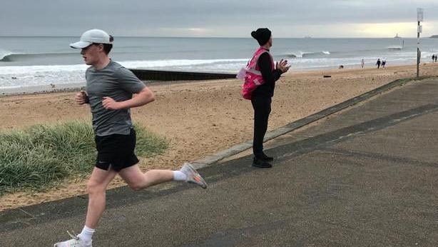 An image of Charles McManus running wearning a grey top and black shorts with the beach and the sea behind 