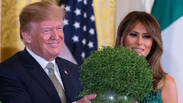 President Donald Trump holding bowl of shamrock, he is smiling and wearing a dark blue suit and gold tie with a white shirt. Beside him is his wife Melania who is wearing a dark green dress.