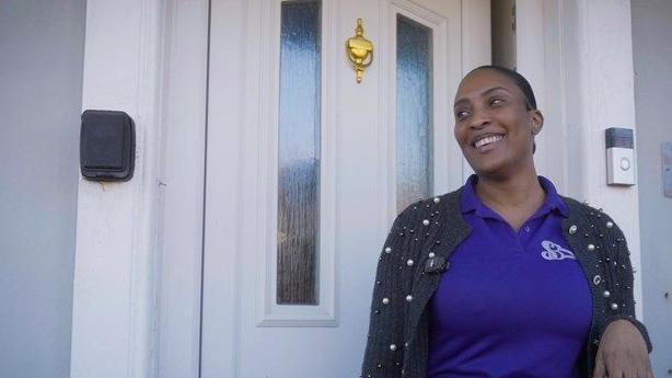 Sasha Beckford stands outside her 1920s home with the front door closed behind her. It has glass panes and a door knocker. She is smiling, looking happy, and is wearing a purple top with a black cardigan.