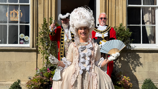 A woman in a Regency inspired dress holding a fan and wearing a white wig. Behind her stands two men in red capes with white ruffles on the front of their outfits.