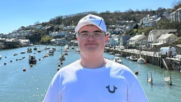 Simon photographed in front of a harbour - there's bright sunshine reflecting off the water and there's lots of fishing boats visible. Simon is wearing a white t shirt and a white baseball cap. He's smiling and looking into the camera. 
