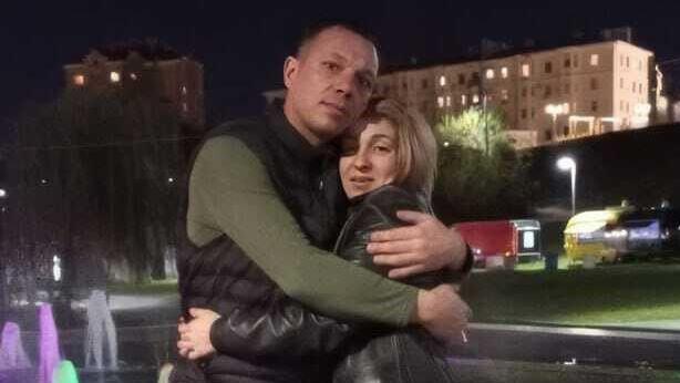 A man in a black top hugs his wife, with blonde hair and a black leather jacket