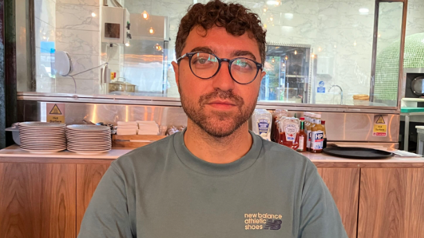 Mehmet Basbaydar sitting in his cafe, smiling at the camera whilst wearing a grey t-shirt and black framed glasses.